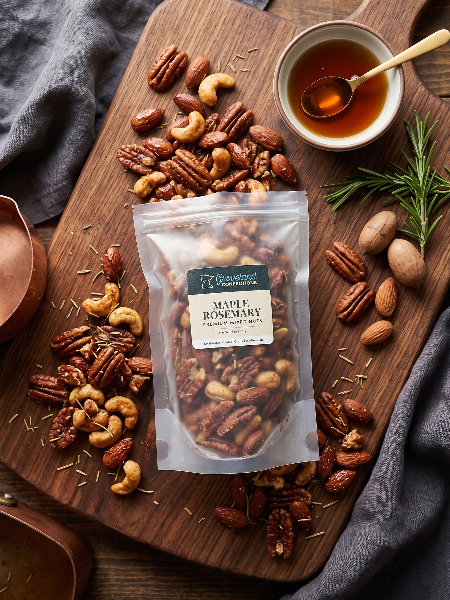 Overhead view of a 7 oz bag of Groveland Confections Maple Rosemary mixed nuts on a walnut board, surrounded by roasted pecans, almonds, and cashews with a bowl of maple syrup and fresh rosemary.